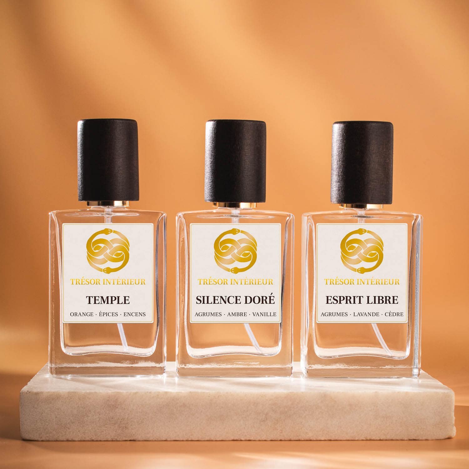 Trois flacons de spray parfum d’intérieur Trésor Intérieur : « Temple », « Esprit Libre » et « Silence Doré » 30ml, sur fond orange.